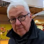 Close-up of an older man with white hair and round black glasses, wearing a dark coat, standing in a shopping mall.