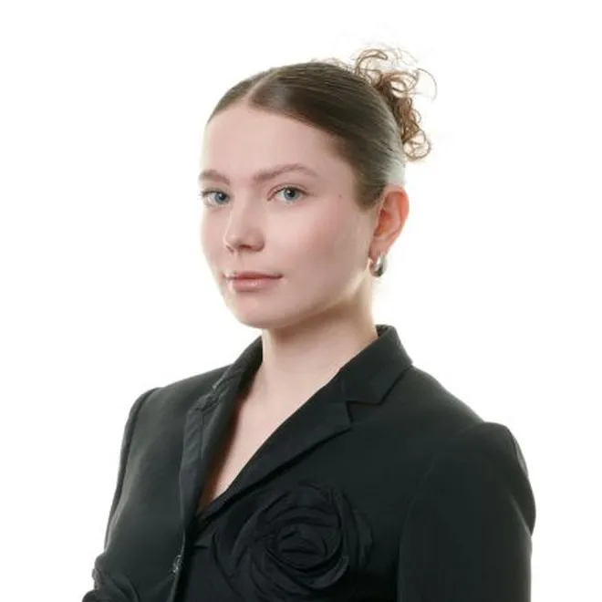 Porträt einer jungen Frau mit heller Haut, die einen schwarzen Blazer mit einer Stoffrose als Verzierung trägt und deren Haare zu einem Dutt zusammengebunden sind, vor weißem Hintergrund.