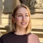 Smiling young woman with short blonde hair and hoop earrings, standing outdoors in front of a stone building with ornate architectural details.