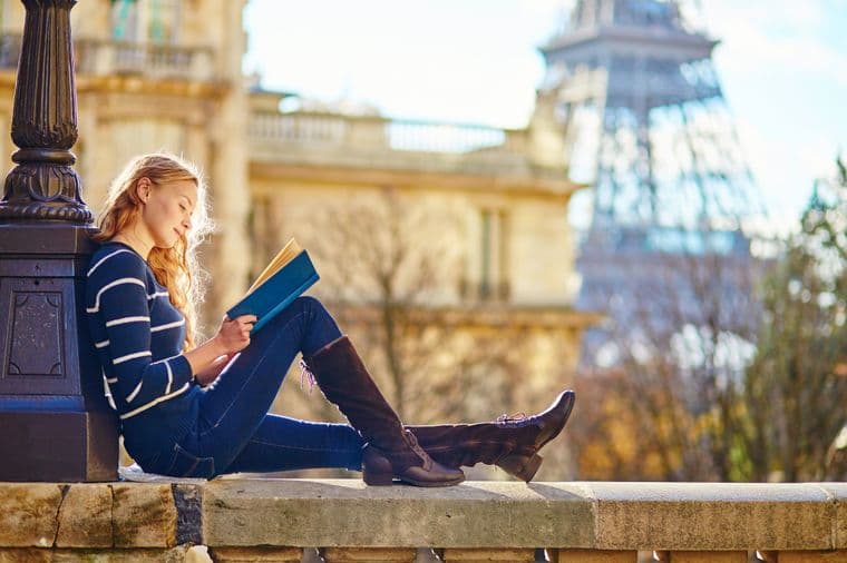 A woman reads a book in Paris. She wears a striped sweater, jeans, and boots. The Eiffel Tower is in the background.