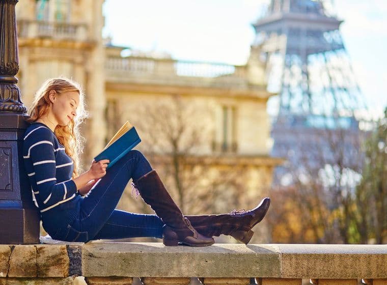 A woman reads a book in Paris. She wears a striped sweater, jeans, and boots. The Eiffel Tower is in the background.