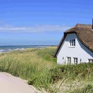 A white cottage with a thatched roof sits beside tall grass dunes overlooking a sandy beach and ocean under a clear blue sky.