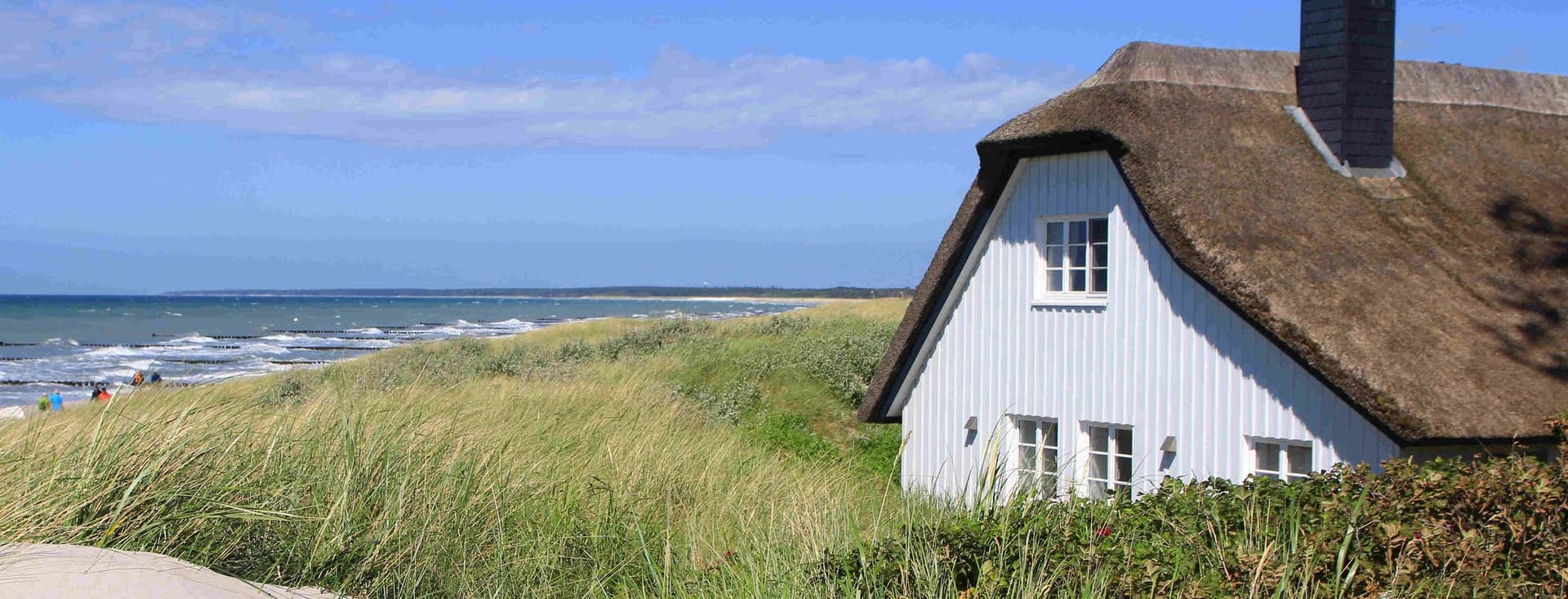 A white cottage with a thatched roof sits beside tall grass dunes overlooking a sandy beach and ocean under a clear blue sky.