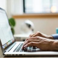 "Person typing on a computer, wearing a checkered shirt, doing real estate marketing."