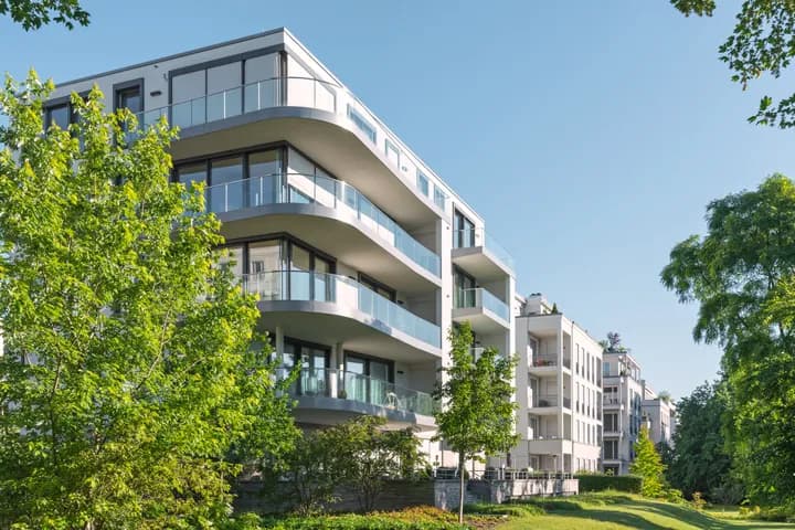 Modern white apartment building with curved glass balconies framed by green trees under a clear blue sky.