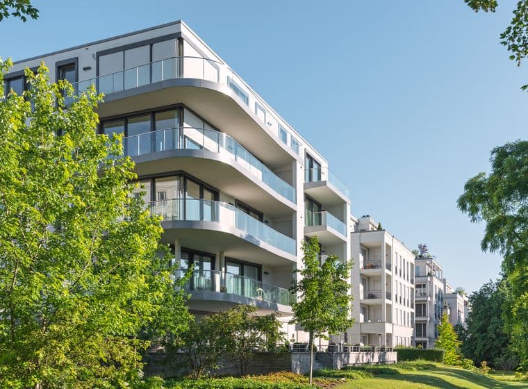 Modern white apartment building with curved glass balconies framed by green trees under a clear blue sky.