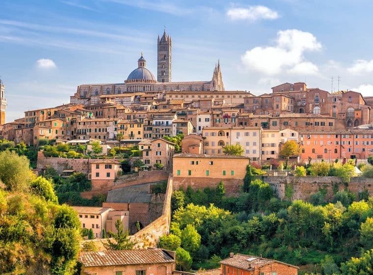 Scenic view of Siena, Italy, showcasing historic buildings and the Siena Cathedral, surrounded by lush greenery under a bright blue sky.