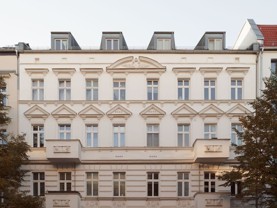 Elegant white neoclassical building facade with ornate details, balconies, and dormer windows, framed by trees on either side.
