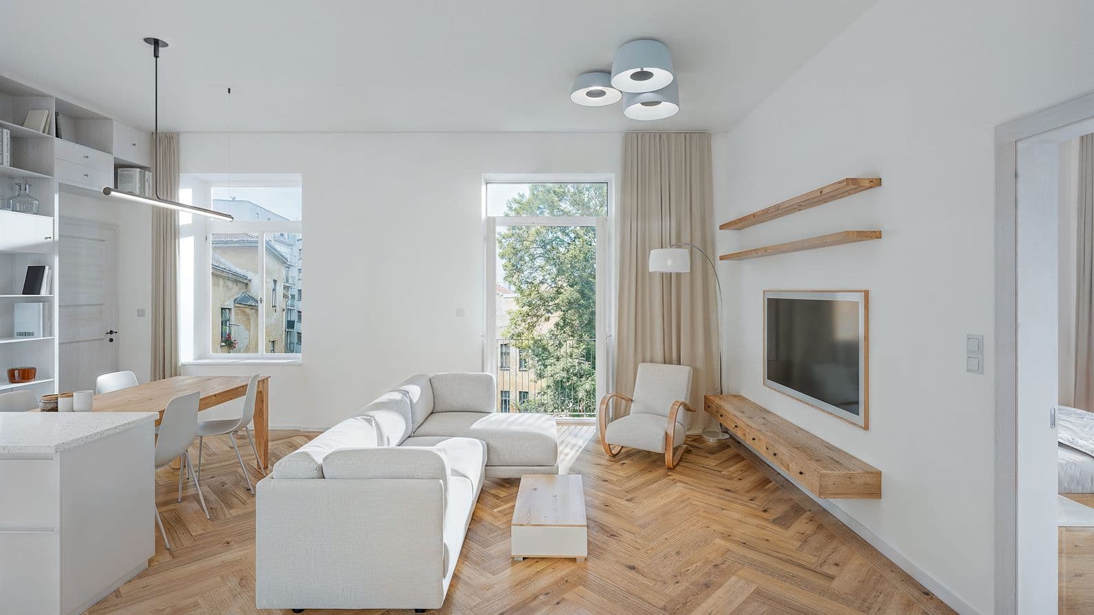 Bright open-plan living room connected to a dining area and kitchen. The space features herringbone wooden flooring, a light sectional sofa, a coffee table, and an armchair. A wall-mounted TV is positioned above a floating wooden cabinet with two wooden shelves. At the rear of the room, a dining table with four chairs sits beside large windows overlooking trees and neighboring buildings. Obrázek vygenerovaný umělou inteligencí