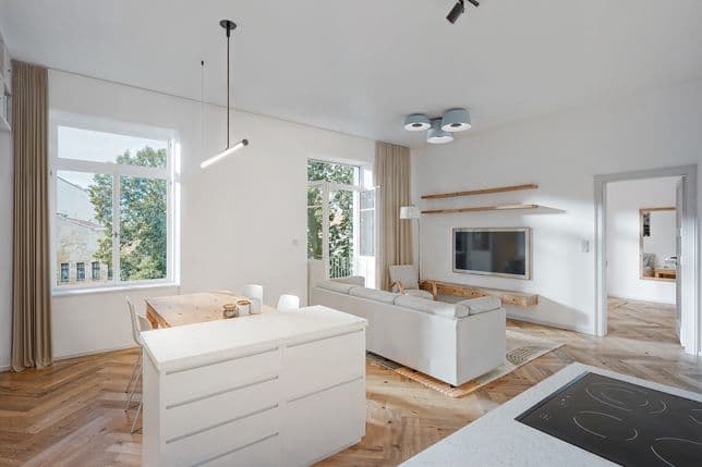 Bright open-plan interior combining the kitchen, dining area, and living room. In the foreground is a white kitchen island with an induction cooktop, next to a wooden dining table with four chairs. The living area features a light-colored sofa, an armchair, a wall-mounted TV above a floating wooden cabinet, and two wooden shelves. Large windows and a balcony door fill the space with natural light and offer views of the surrounding greenery. Obrázek vygenerovaný umělou inteligencí