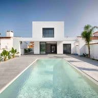 Vista d'una casa moderna blanca amb piscina a l'entrada, jardí, palmeres al voltant i cels blaus.