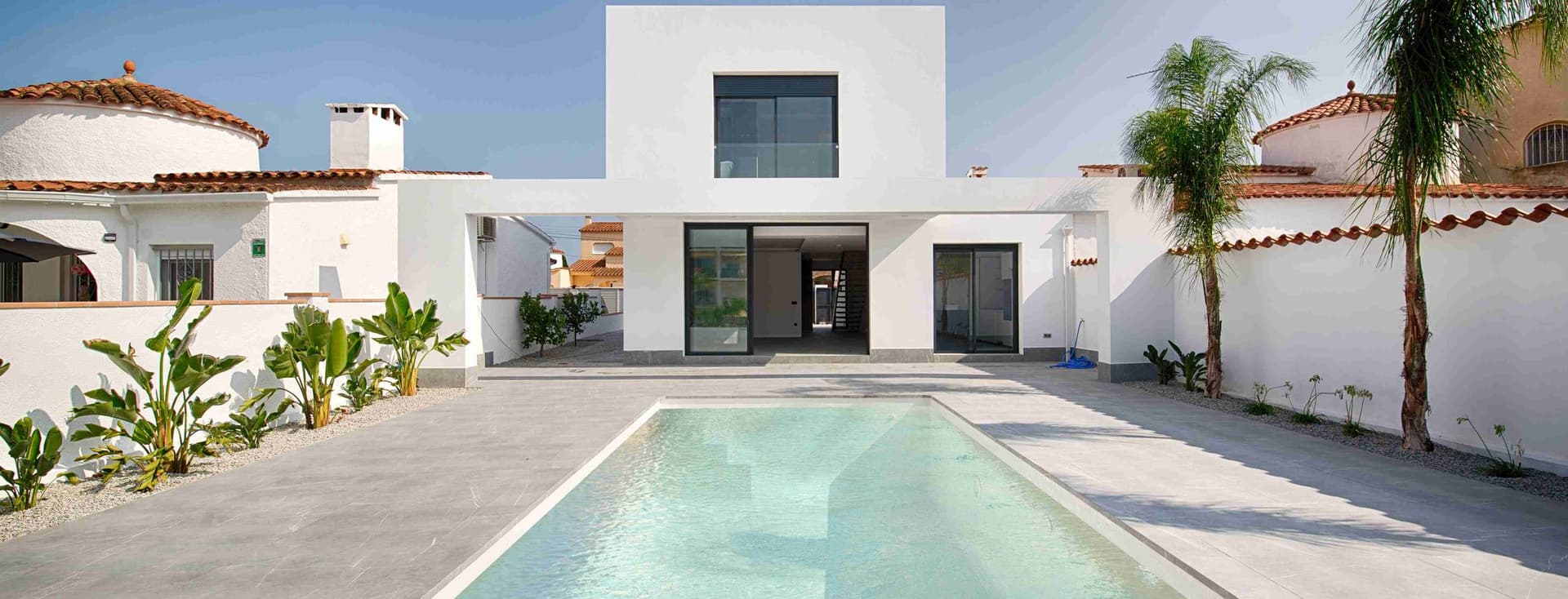 Vista d'una casa moderna blanca amb piscina a l'entrada, jardí, palmeres al voltant i cels blaus.