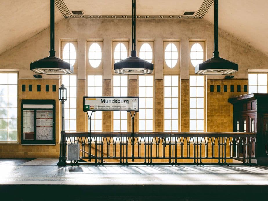 Sunlit train station platform with vintage lamps, arched windows, and a sign reading "Mundsburg." Elegant architecture and warm tones.