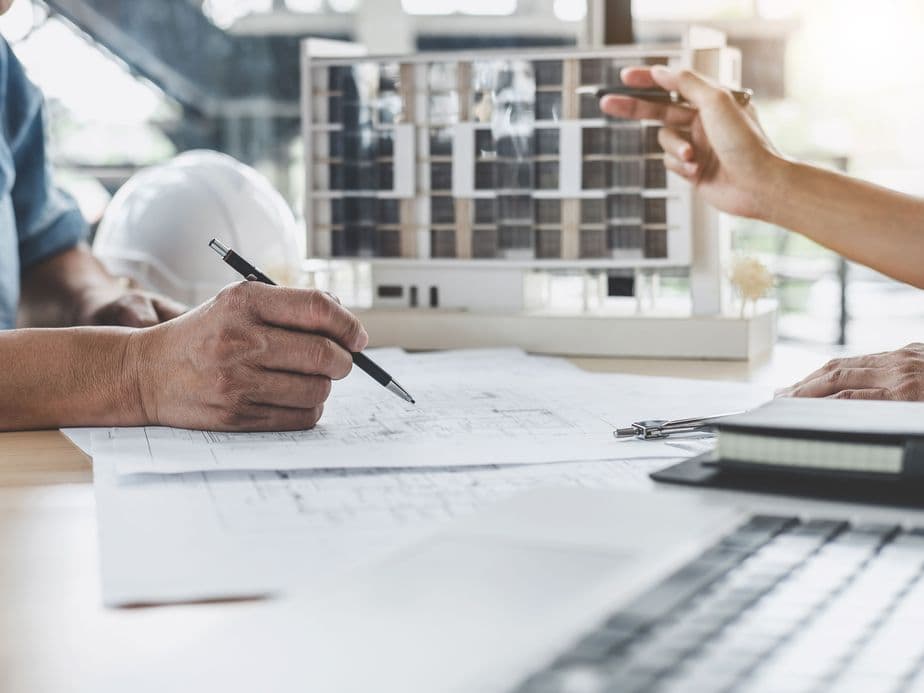 Two people discuss architectural plans at a desk with blueprints, a laptop, and a building model. One points while the other takes notes.
