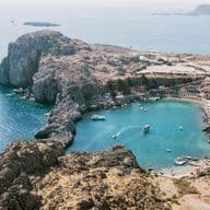 Aerial view of a rocky coastline with a small bay, clear turquoise water, boats, and a winding road on a sunny day.