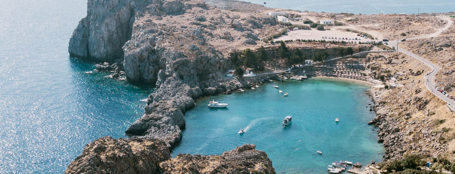 Aerial view of a rocky coastline with a small bay, clear turquoise water, boats, and a winding road on a sunny day.