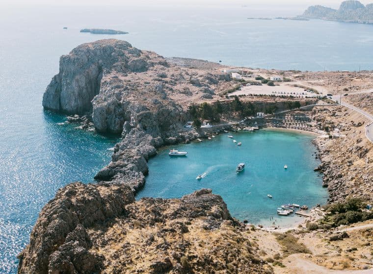 Aerial view of a rocky coastline with a small bay, clear turquoise water, boats, and a winding road on a sunny day.