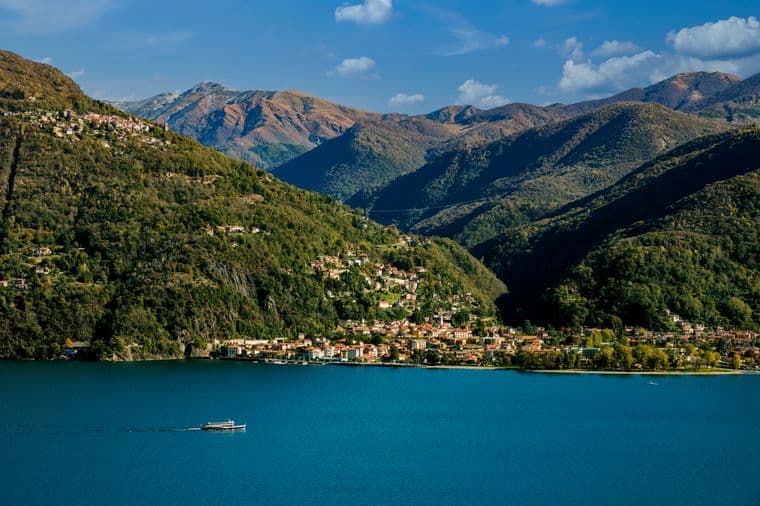 Veduta panoramica di Varese dal lago: il profilo della città immerso nel verde, con colline sullo sfondo e riflessi sull’acqua in una giornata luminosa.