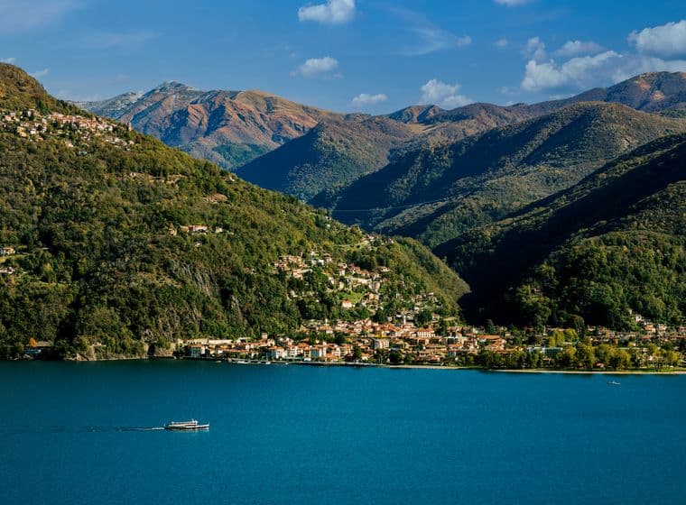 Veduta panoramica di Varese dal lago: il profilo della città immerso nel verde, con colline sullo sfondo e riflessi sull’acqua in una giornata luminosa.