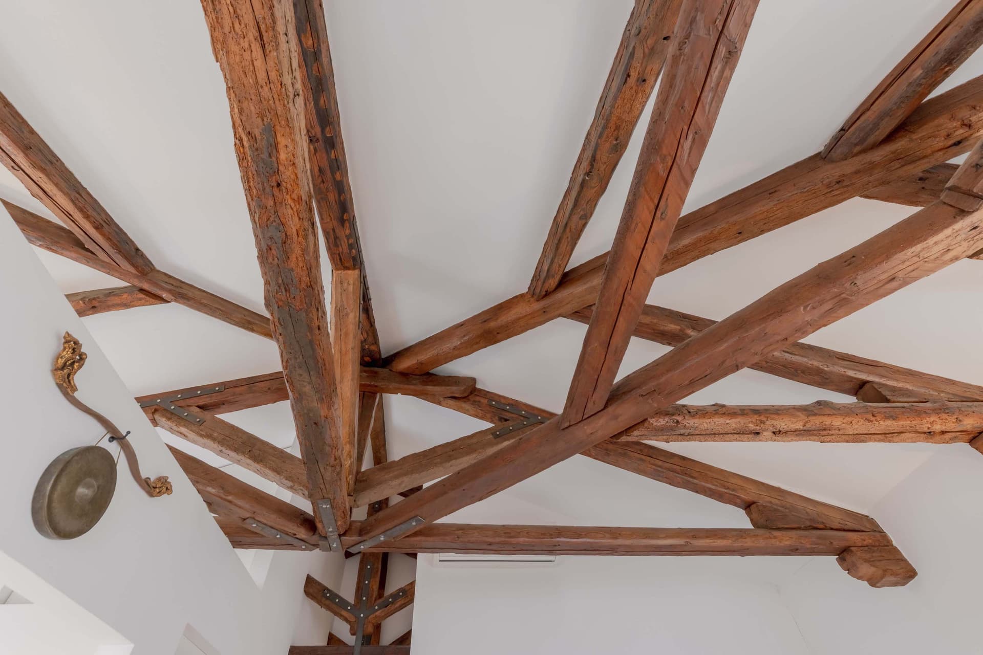 The ceiling of a modern interior featuring an exposed structure of antique wooden beams. The beams are arranged in an interwoven pattern and made of rustic wood in a reddish-brown hue, showcasing natural characteristics and signs of aging. The beams are supported by metallic elements. The ceiling is white, creating a sharp contrast with the warm tones of the wood. A lamp or decorative element is subtly visible hanging on the side wall.