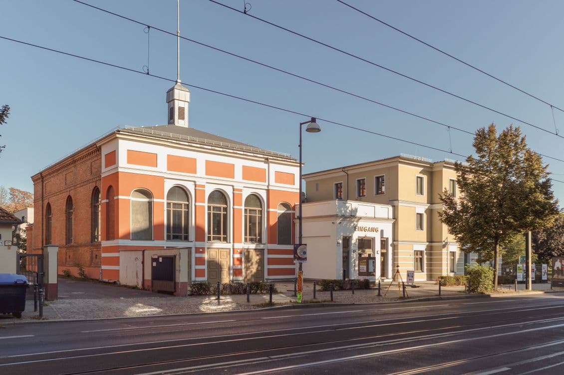 Street view of a historic red and beige building with arched windows, next to a modern beige structure, under clear blue sky.