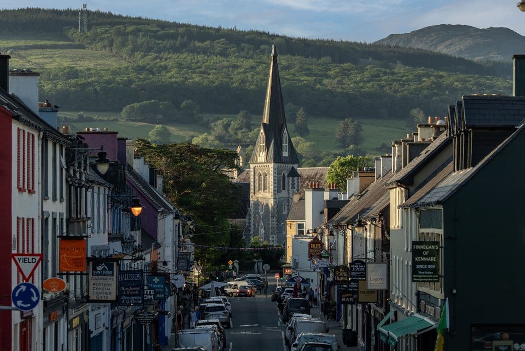Charming main street of Kenmare, Ireland, with colorful shops and businesses lining both sides of the street, a church spire visible in the center, and rolling green hills and mountains in the background under a blue sky
