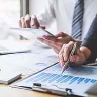 Two people reviewing financial charts and graphs on paper and a tablet at a desk, with a laptop and sticky notes nearby.