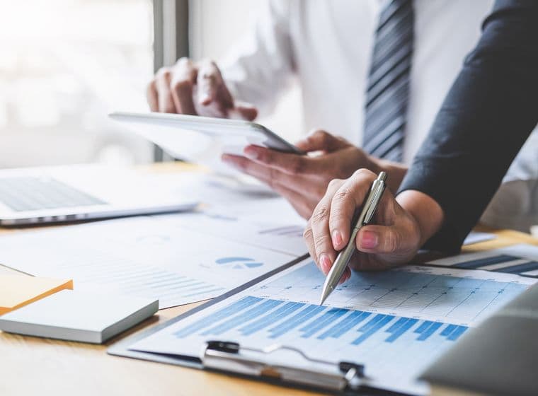 Two people reviewing financial charts and graphs on paper and a tablet at a desk, with a laptop and sticky notes nearby.