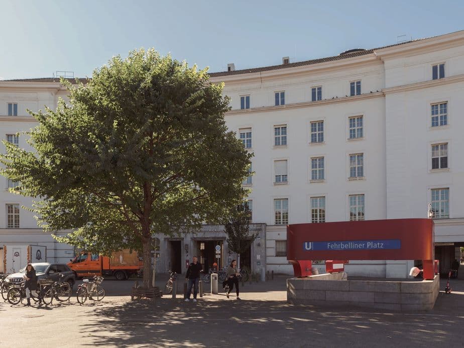 A curved white building with multiple windows, a large tree, bicycles, and people near the Fehrbelliner Platz U-Bahn station entrance.