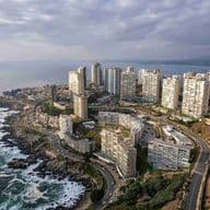 Aerial view of a coastal city with white high-rise buildings, winding roads, and rocky shoreline under a cloudy sky.