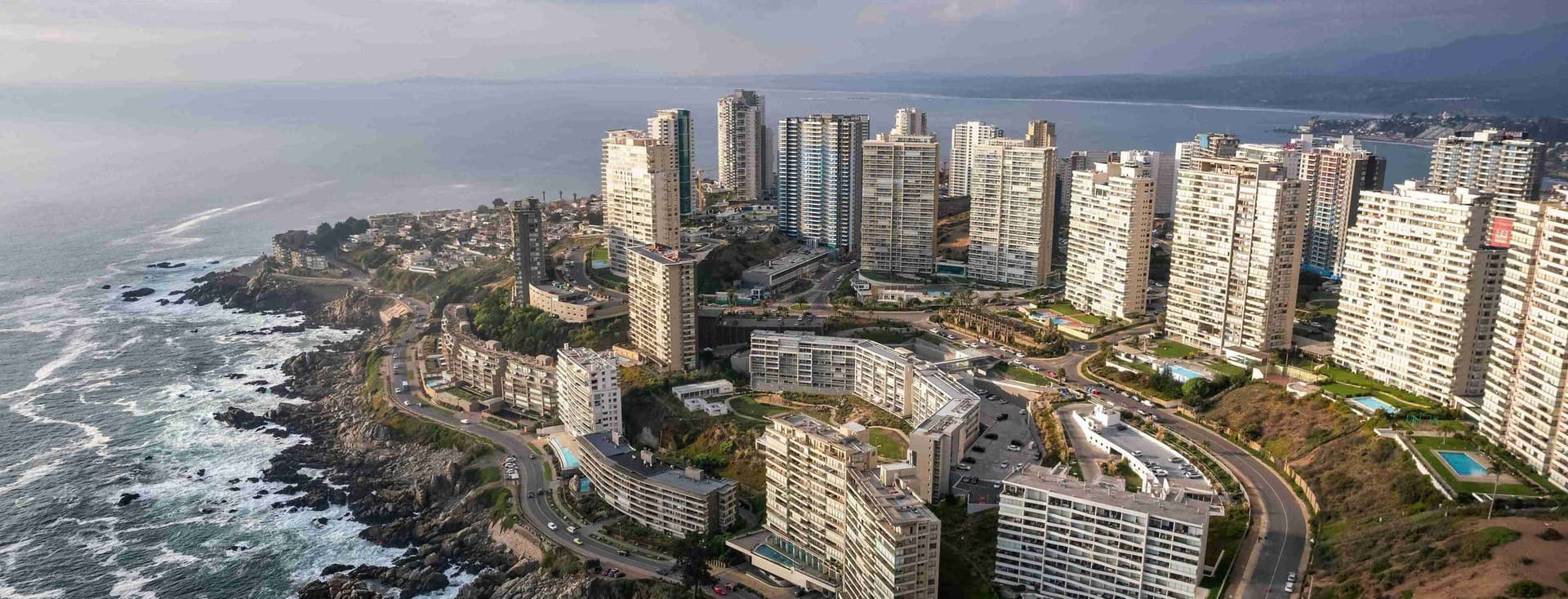 Aerial view of a coastal city with white high-rise buildings, winding roads, and rocky shoreline under a cloudy sky.