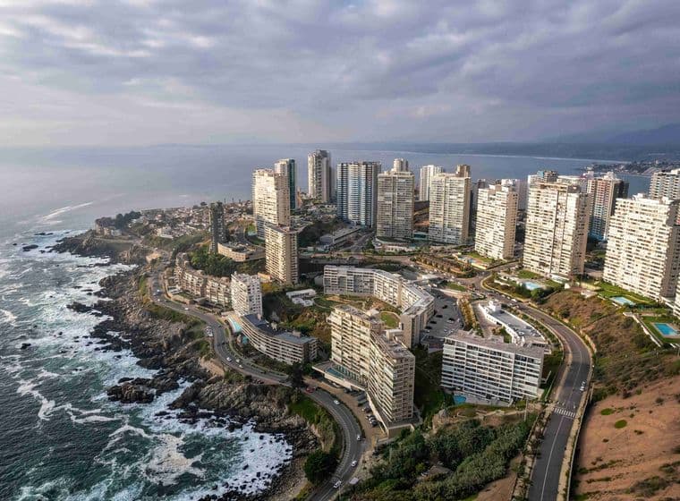 Vista aérea de una ciudad costera con edificios blancos de gran altura, carreteras sinuosas y costa rocosa bajo un cielo nublado.