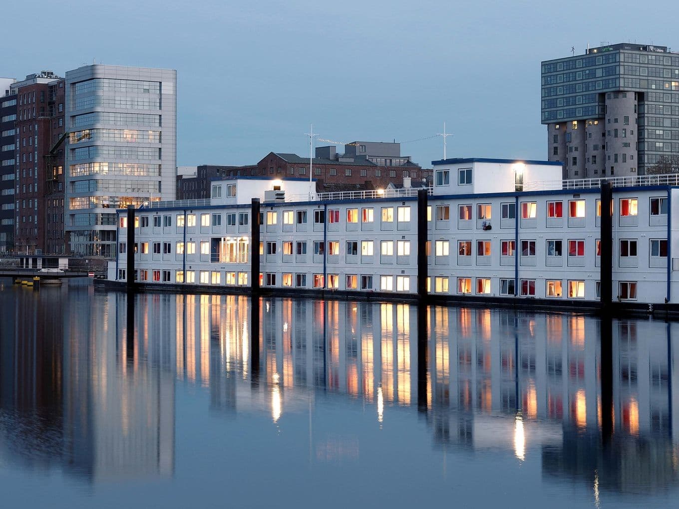 Modern buildings on calm water at dusk, in Harburg, with a clear sky in the background.
