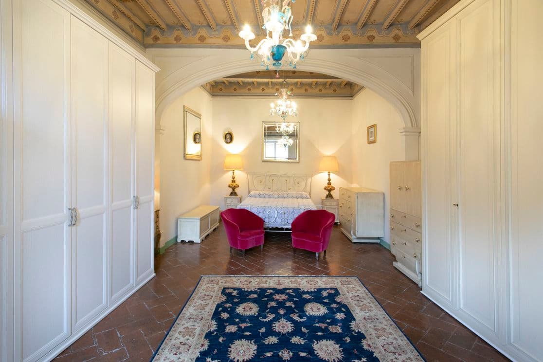 Spacious bedroom with ornate ceiling, chandelier, double bed, two red chairs, and a blue patterned rug on tiled floor.