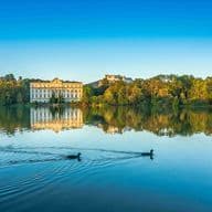 Evening view of Schloss Hellbrunn, a majestic baroque palace in South Salzburg, perfectly reflected in its calm palace pond. Two ducks glide across the foreground waters, while the golden evening light illuminates the palace's yellow facade and the surrounding autumn trees. The historic estate exemplifies the prestigious real estate offerings in Salzburg's southern district.