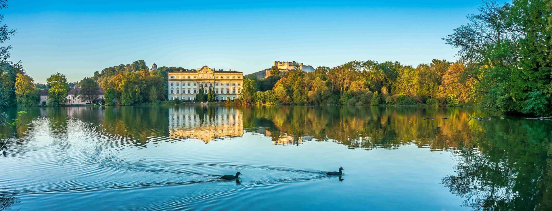 Schloss Hellbrunn spiegelt sich im Teich bei Sonnenuntergang, mit schwimmenden Enten im Vordergrund und Festung Hohensalzburg im Hintergrund unter blauem Himmel.