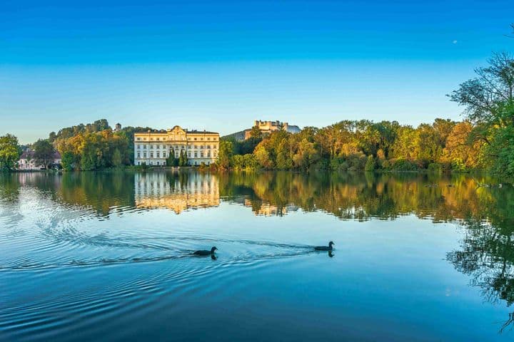 Evening view of Schloss Hellbrunn, a majestic baroque palace in South Salzburg, perfectly reflected in its calm palace pond. Two ducks glide across the foreground waters, while the golden evening light illuminates the palace's yellow facade and the surrounding autumn trees. The historic estate exemplifies the prestigious real estate offerings in Salzburg's southern district.