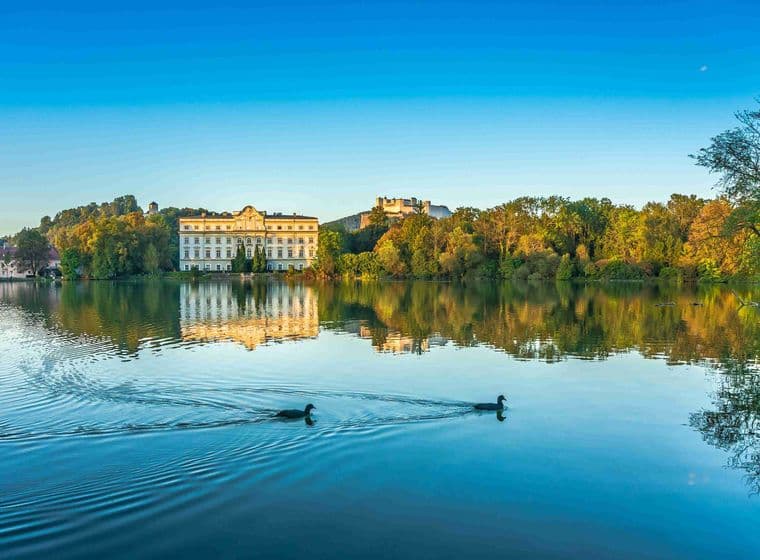 Schloss Hellbrunn spiegelt sich im Teich bei Sonnenuntergang, mit schwimmenden Enten im Vordergrund und Festung Hohensalzburg im Hintergrund unter blauem Himmel.