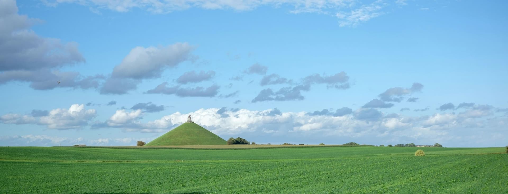 Groen veld met een kegelvormige heuvel op de achtergrond onder een blauwe lucht met witte wolken.