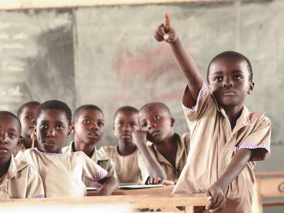 A group of young students in uniforms sit at desks in a classroom. One child raises their hand. A chalkboard is visible in the background.