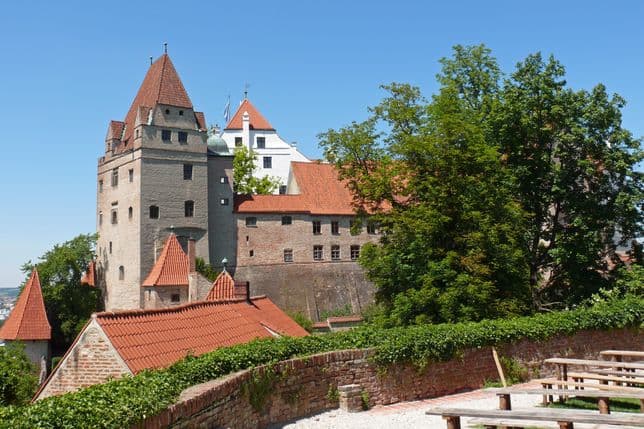 Historic castle with red-tiled roofs and stone walls, surrounded by greenery under a clear blue sky. Wooden benches in the foreground.