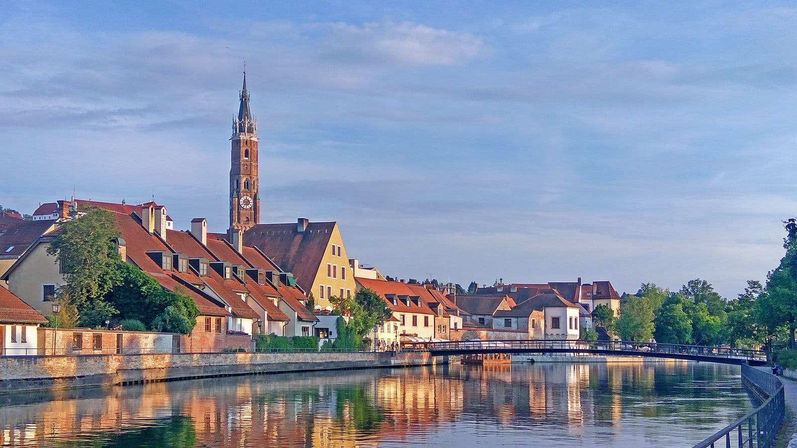 A scenic view of a riverside town with colorful houses, a tall clock tower, and reflections on the calm water under a clear blue sky.