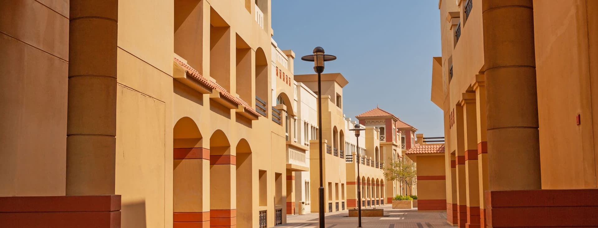 A sunny courtyard with Mediterranean-style architecture, featuring arches, columns, and a lamp post on a patterned stone pathway.