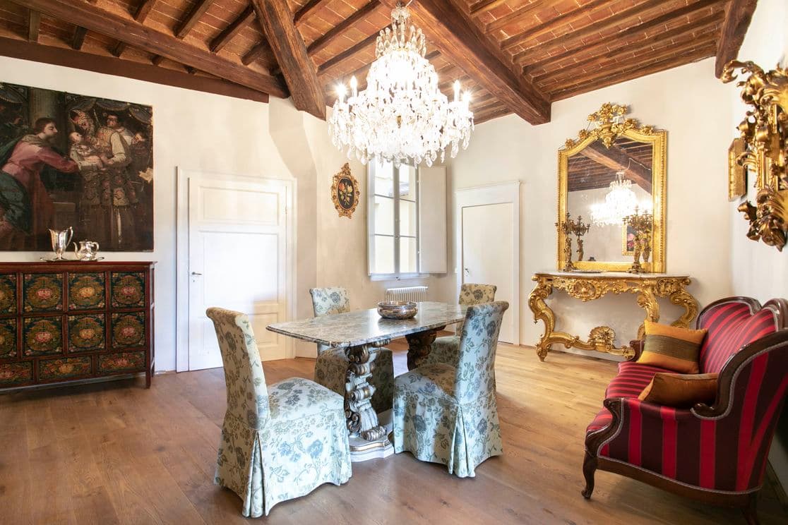 Elegant dining room with a marble table, floral chairs, ornate chandelier, gold-framed mirror, and vintage decor on wooden floors and beamed ceiling.