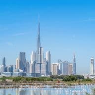 Dubai skyline with the Burj Khalifa towering over other skyscrapers, set against a clear blue sky and a foreground of water and greenery.