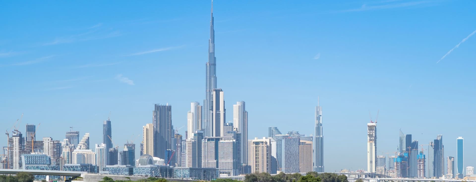 Dubai skyline with the Burj Khalifa towering over other skyscrapers, set against a clear blue sky and a foreground of water and greenery.