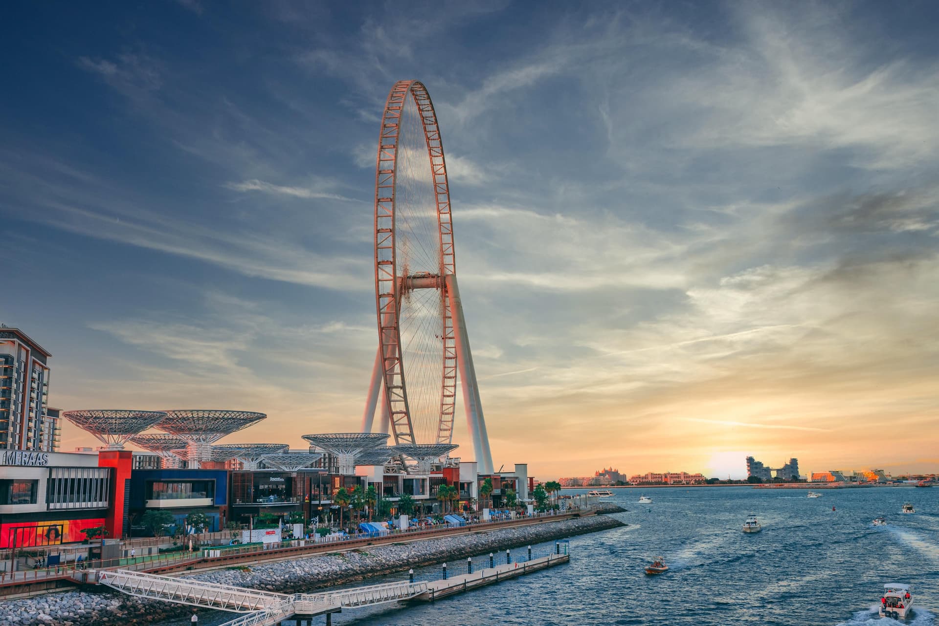 Das Ain Riesenrad von Dubai ragt hoch vor einem Sonnenuntergangshimmel. Boote säumen das Wasser in der Nähe einer Uferpromenade mit Geschäften und Restaurants.