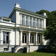 White neoclassical building with tall decorative columns, arched windows, and balconies, beside green trees under a clear blue sky.