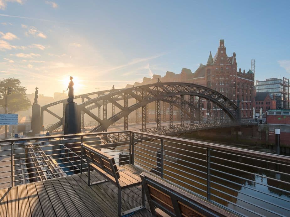 Sunrise over a historic iron bridge and brick buildings by a canal, with benches on a wooden deck in the foreground.