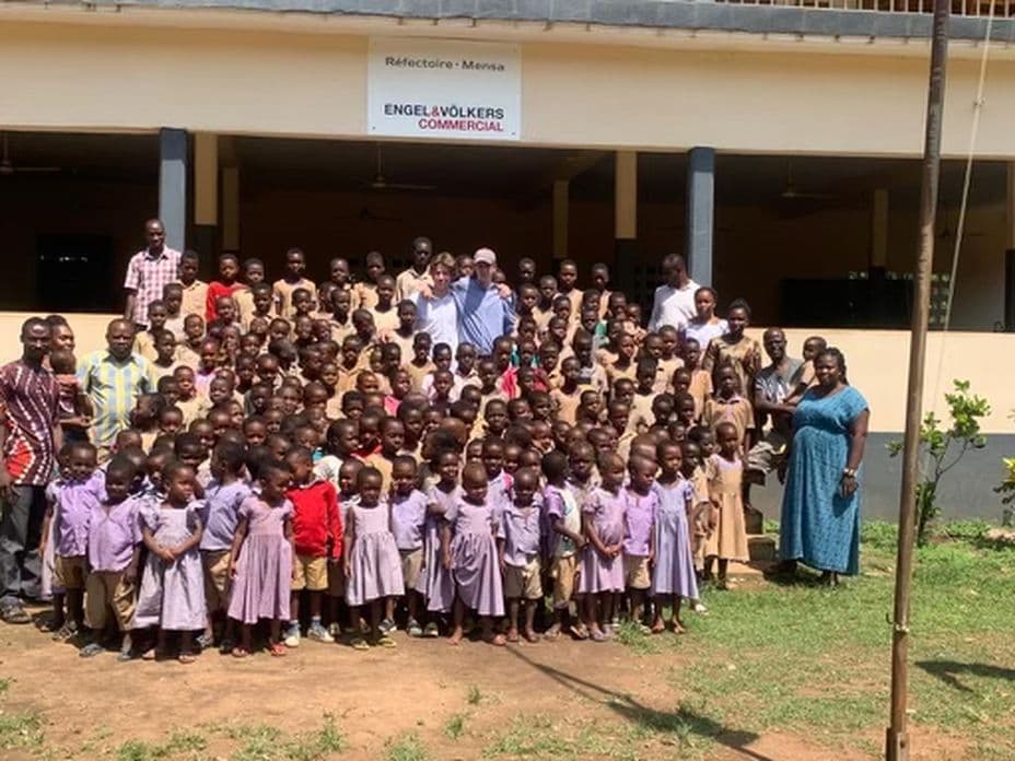 A large group of children and adults pose together in front of a school building with a sign above the entrance.